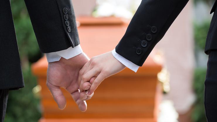 Hände von zwei Personen in eleganter Kleidung, die sich über einem Holzsarg halten.