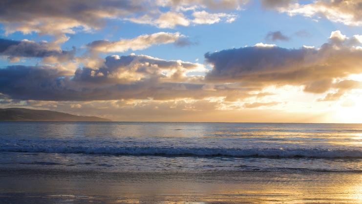 Sonnenuntergang über dem Meer, sanfte Wellen brechen am ruhigen Strand, Wolken reflektieren das Licht am Himmel.