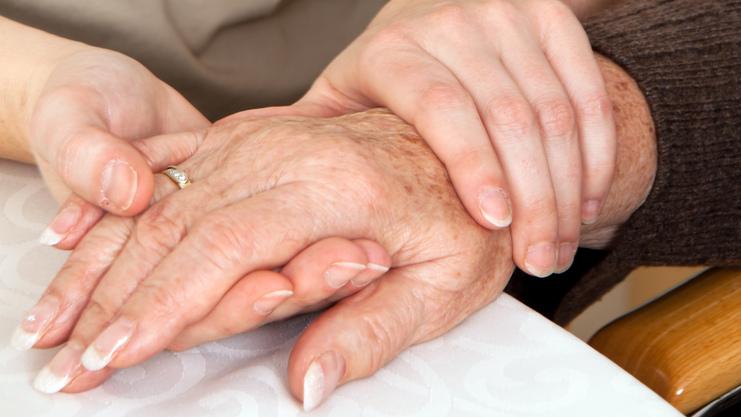 Zwei Hände, eine ältere und eine jüngere, liegen auf einem Tisch, wobei die jüngere Hand die ältere sanft umschließt.