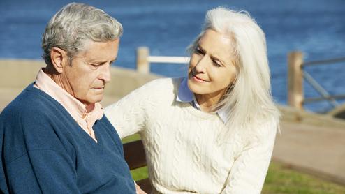 Ein älteres Paar sitzt auf einer Bank am Wasser, die Frau hält den Blick des Mannes sanft und einfühlsam.