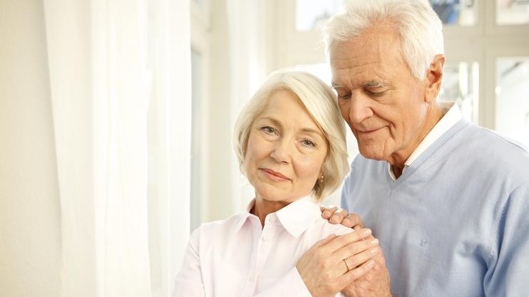 Ein älteres Paar steht nah beieinander, der Mann umarmt die Frau sanft, beide lächeln in eine ruhige Umgebung.